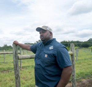 Davon wears a blue shirt and tan baseball cape he looks to the side of the camera and his hand is resting on a fence post. 
